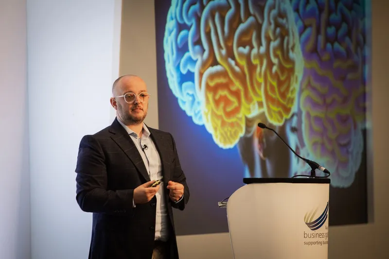 Martin Broadhurst presenting on AI at a conference, a large brain visualisation on the screen behind him